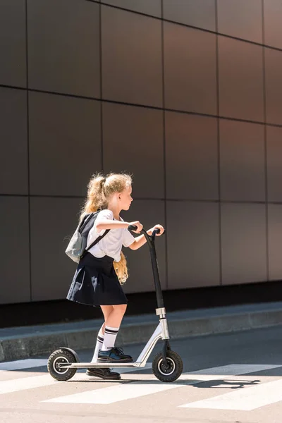 Вид сбоку восхитительной маленькой школьницы с рюкзаком, катающейся на скутере по улице — стоковое фото