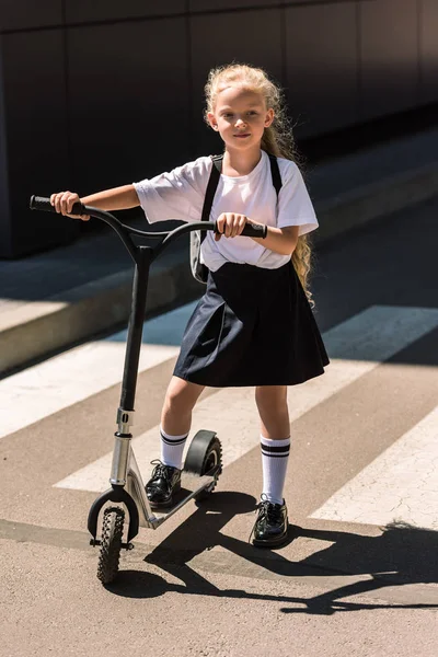 Full length view of adorable little schoolgirl with backpack riding scooter on street — Stock Photo