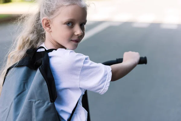 Adorable escolar con mochila a caballo scooter y mirando a la cámara - foto de stock