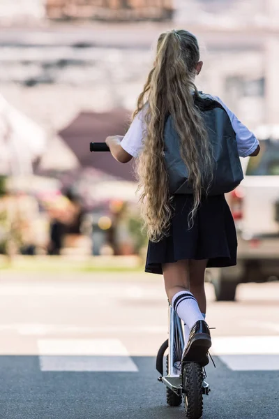 Back view of little schoolgirl with long curly hair riding scooter on street — Stock Photo