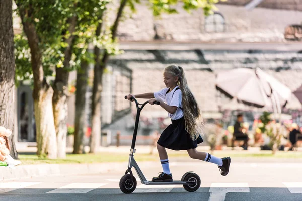 Side view of adorable happy schoolchild with backpack riding scooter on street — Stock Photo