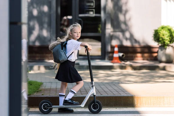 Entzückendes Schulmädchen mit Rucksack und Teddybär, das Roller fährt und auf der Straße in die Kamera schaut — Stockfoto