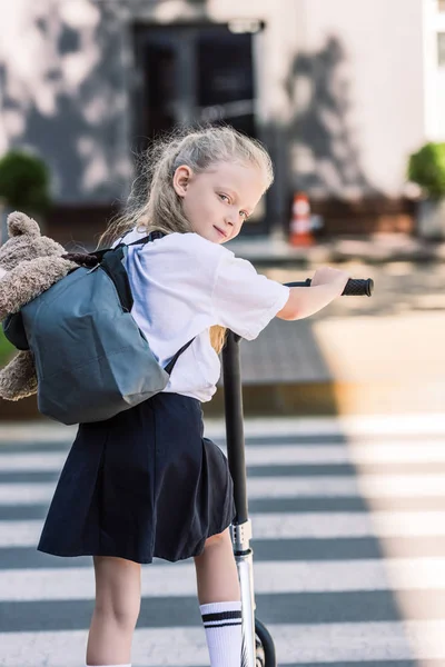 Back view of adorable little schoolgirl with backpack and teddy bear riding scooter and looking at camera — Stock Photo