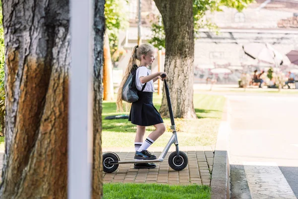 Side view of cute little schoolgirl with backpack riding scooter on street — Stock Photo