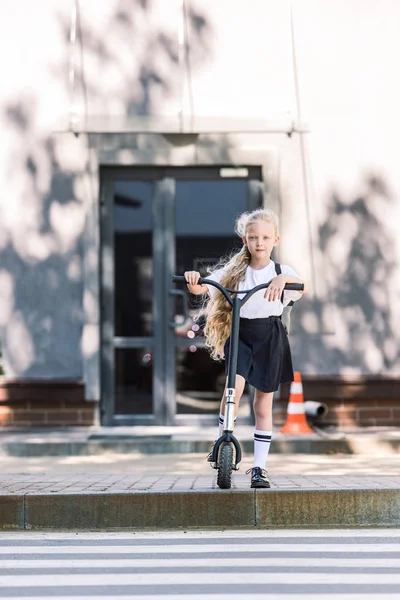 Adorable schoolgirl riding scooter and looking at camera on street — Stock Photo