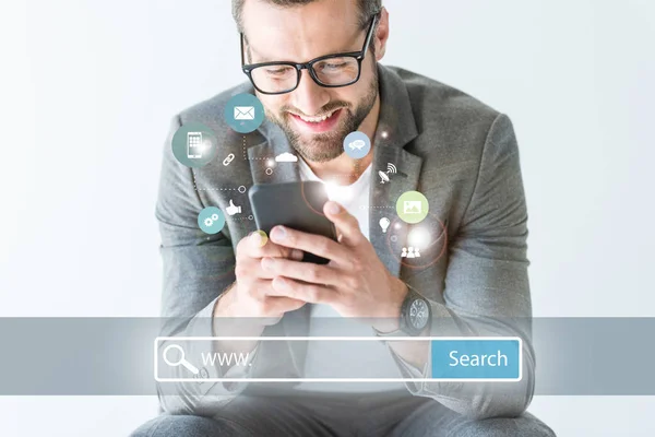 Усміхнений розробник SEO у сірому костюмі з використанням смартфона, ізольований на білому з панеллю пошуку веб-сайтів та іконками — стокове фото