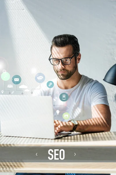 Bearded developer working with laptop with SEO icons — Stock Photo