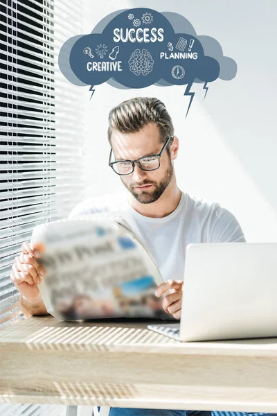 Handsome businessman reading newspaper at workplace with laptop, with success, planning, creative signs — Stock Photo