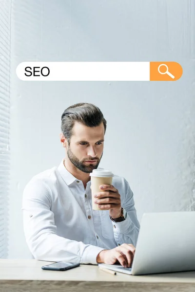 Concentrated developer holding coffee to go while working with laptop with SEO search bar — Stock Photo