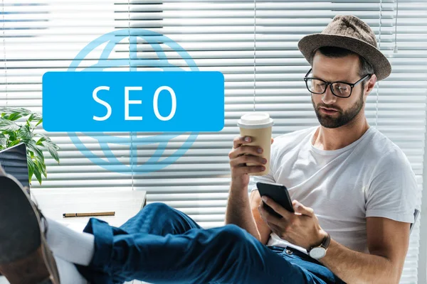 Bearded developer in hat holding coffee to go and using smartphone with SEO sign — Stock Photo