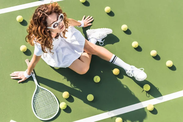 Overhead view of fashionable tennis player in white sportswear and sunglasses resting on tennis court with balls and racket near by — Stock Photo