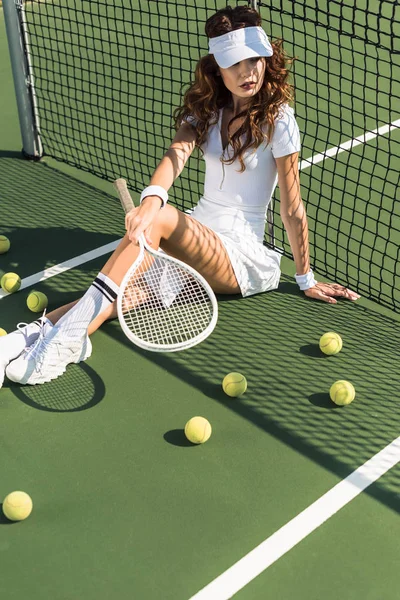 Elegante tenista en ropa deportiva blanca con raqueta de tenis sentado en la red en la pista de tenis - foto de stock