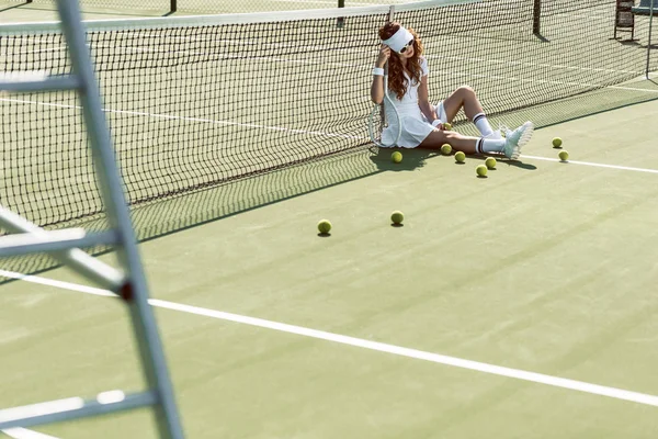 Elegante jugadora de tenis en gafas de sol descansando cerca de la red en la cancha de tenis con equipo cerca — Stock Photo