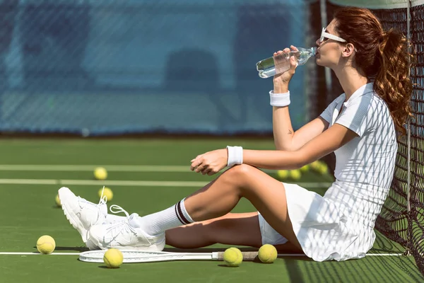Hermosa tenista en uniforme de tenis blanco y gafas de sol agua potable mientras descansa en la cancha con raqueta y pelotas - foto de stock