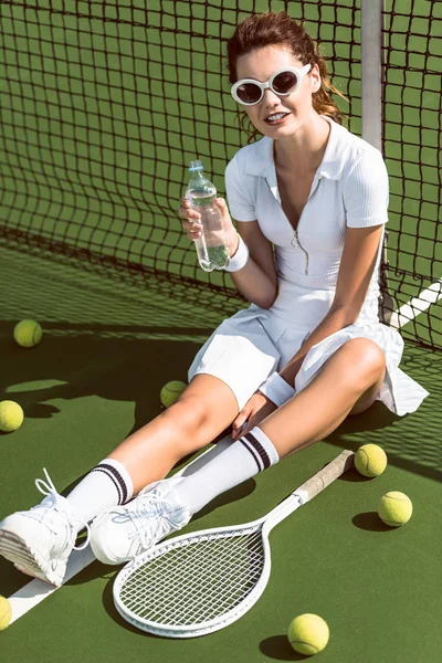 Beautiful tennis player in white tennis uniform and sunglasses with water bottle resting on court with racket and balls — Stock Photo