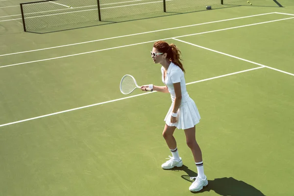 Schöne Frau in weißer Sportbekleidung und Sonnenbrille spielt Tennis auf dem Platz — Stockfoto