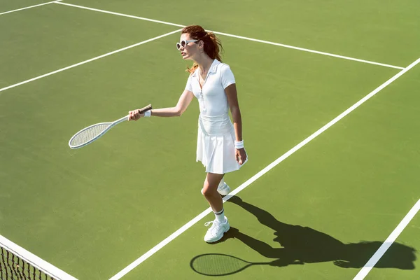 Schöne Frau in weißer Sportbekleidung und Sonnenbrille spielt Tennis auf dem Platz — Stockfoto