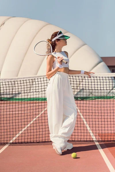 Hermosa mujer en ropa blanca y gorra con raqueta de tenis posando en la red de tenis en la cancha - foto de stock