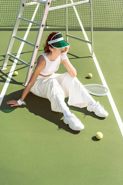 Young woman in fashionable white clothing and cap sitting on tennis court with racket and balls — Stock Photo