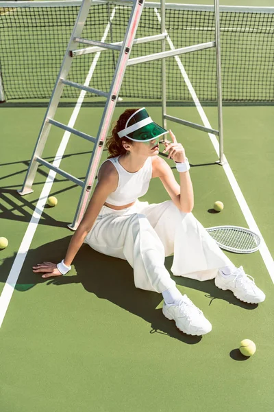 Jeune femme en vêtements blancs à la mode et casquette assise sur un court de tennis avec raquette et balles — Photo de stock