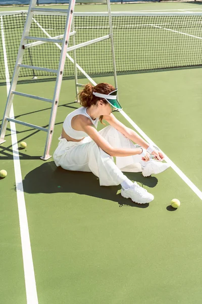 Vista lateral da jovem mulher em roupas brancas elegantes e boné amarrar atacadores no campo de ténis com raquete e bolas — Fotografia de Stock