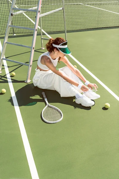 Mujer joven con ropa blanca elegante y gorra atando cordones de zapatos en la cancha de tenis con raqueta y pelotas - foto de stock