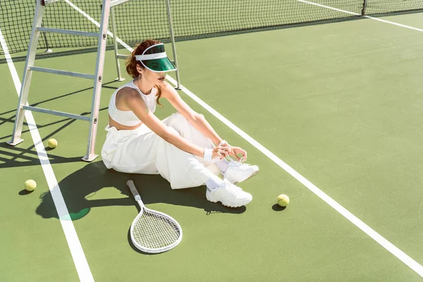 Mujer joven con ropa blanca elegante y gorra atando cordones de zapatos en la cancha de tenis con raqueta y pelotas - foto de stock