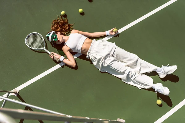 Hochwinkelaufnahme einer stilvollen Frau in weißer Kleidung und Mütze, die mit Schläger auf Tennisplatz liegend mit Schläger liegt — Stockfoto