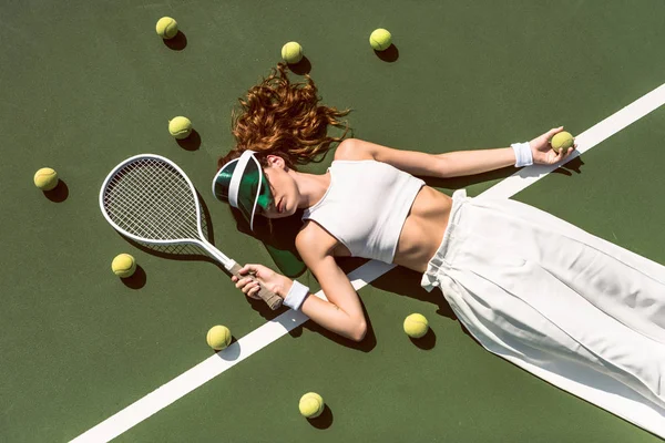 Над головой вид стильной женщины в белой одежде и кепке лежащей с ракеткой на теннисном корте с ракеткой — стоковое фото