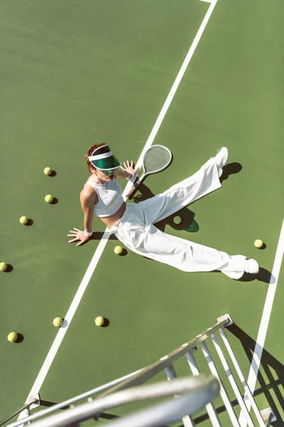 Високий кут зору красивої жінки в стильному білому одязі, що сидить на тенісному корті з м'ячами і ракеткою навколо — стокове фото