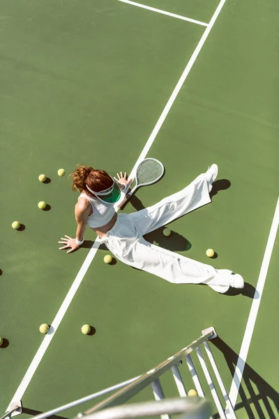 Vista de ángulo alto de la hermosa mujer en ropa blanca con estilo sentado en la cancha de tenis con pelotas y raqueta alrededor - foto de stock