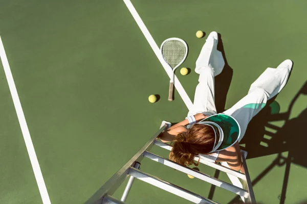 Над головой вид женщины в стильной белой одежде и кепке, позирующей на кресле судьи на теннисном корте — стоковое фото