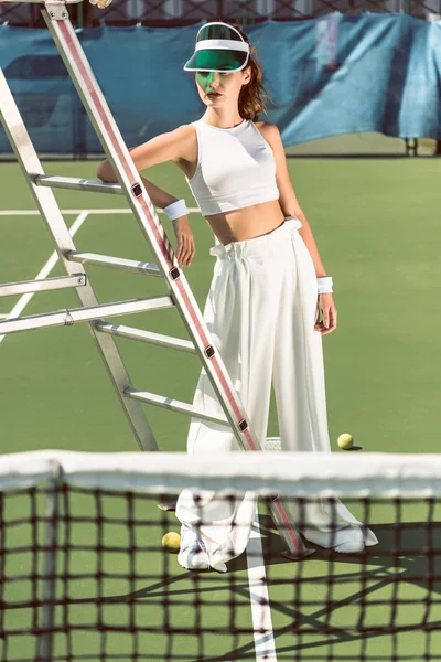 Молода жінка в стильному білому одязі і шапка позує біля арбітражного крісла на тенісному корті — стокове фото