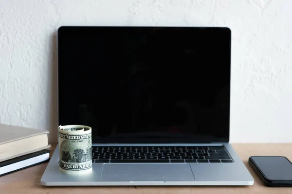 Portátil con pantalla en blanco, teléfono inteligente, billetes en dólares laminados y libros en la mesa - foto de stock