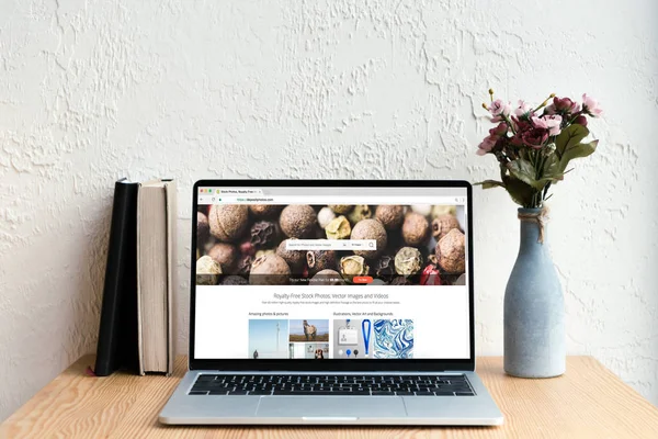 Laptop com site de fotos depositadas na tela, livros e flores em vaso na mesa de madeira — Fotografia de Stock