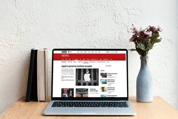 Laptop with bbc news website on screen, books and flowers in vase on wooden table — Stock Photo