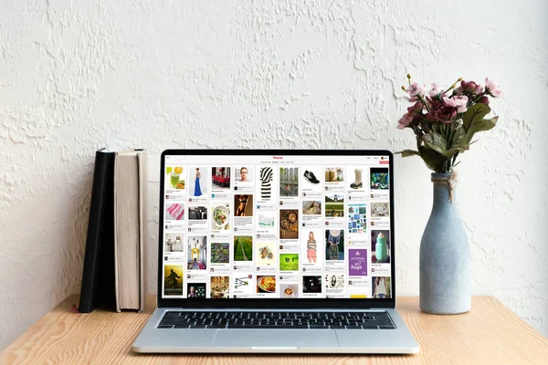 Laptop with pinterest website on screen, books and flowers in vase on wooden table — Stock Photo