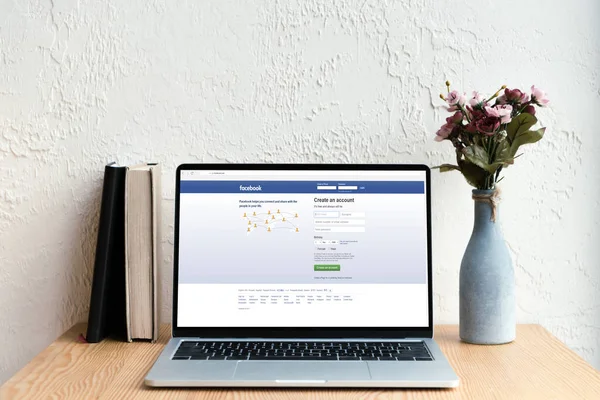 Portátil con página web de facebook en pantalla, libros y flores en jarrón sobre mesa de madera - foto de stock