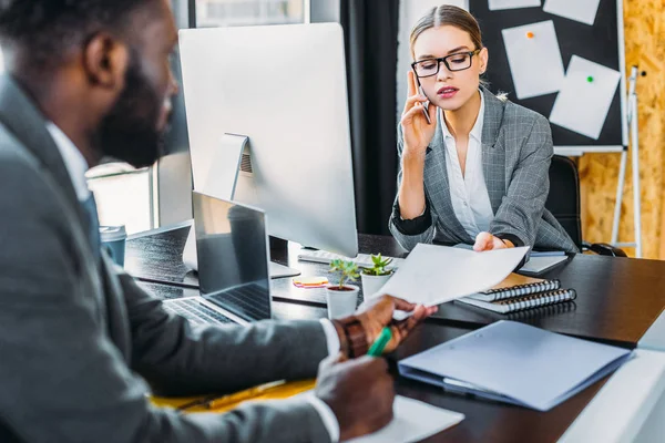 African american businessman giving documents to businesswoman talking by smartphone — Stock Photo