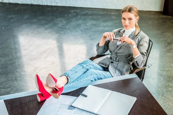 Hochwinkelaufnahme einer Geschäftsfrau, die mit Beinen auf einem Tisch im Büro sitzt und in die Kamera schaut — Stockfoto