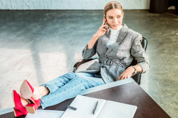 Vista de ángulo alto de la mujer de negocios sentada con las piernas en la mesa en la oficina y hablando por teléfono inteligente - foto de stock