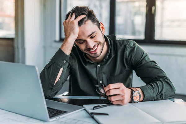 Hombre de negocios sonriente sentado en la mesa en la oficina y sosteniendo gafas - foto de stock