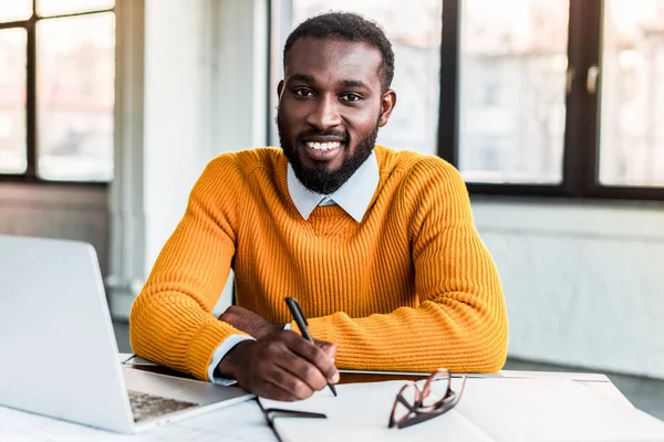 Улыбающийся африканский бизнесмен, держащий ручку и смотрящий на камеру в офисе — стоковое фото