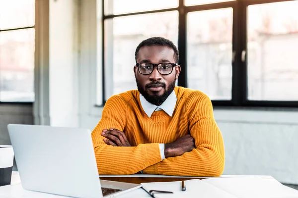 Afrikanischer amerikanischer Geschäftsmann sitzt mit verschränkten Armen im Büro und blickt in die Kamera — Stockfoto