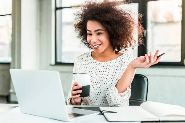 Heureuse femme d'affaires afro-américaine tenant une tasse de café et regardant ordinateur portable au bureau — Photo de stock