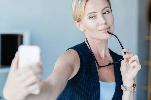 Bella donna d'affari adulta con occhiali prendere selfie su smartphone in ufficio — Foto stock