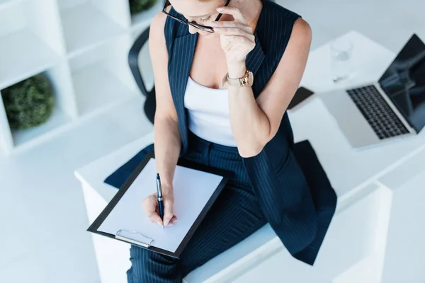 Hochwinkelaufnahme einer Geschäftsfrau, die in Klemmbrett auf Tisch mit Laptop im Büro schreibt — Stockfoto