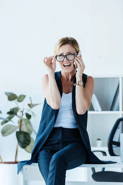 Femme d'affaires émotionnelle touchant lunettes et parler sur smartphone au bureau — Photo de stock