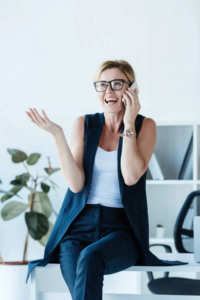 Mujer de negocios sonriente en gafas gestos a mano y hablando en el teléfono inteligente en la oficina - foto de stock