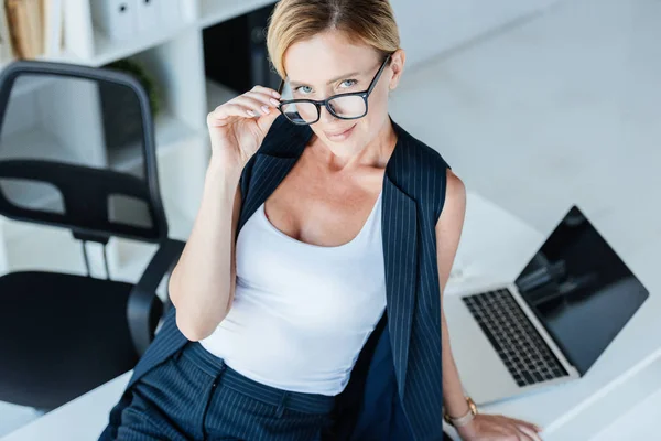 Vista ad alto angolo di attraente donna d'affari che regola gli occhiali e guarda la fotocamera sul tavolo con computer portatile in ufficio — Foto stock